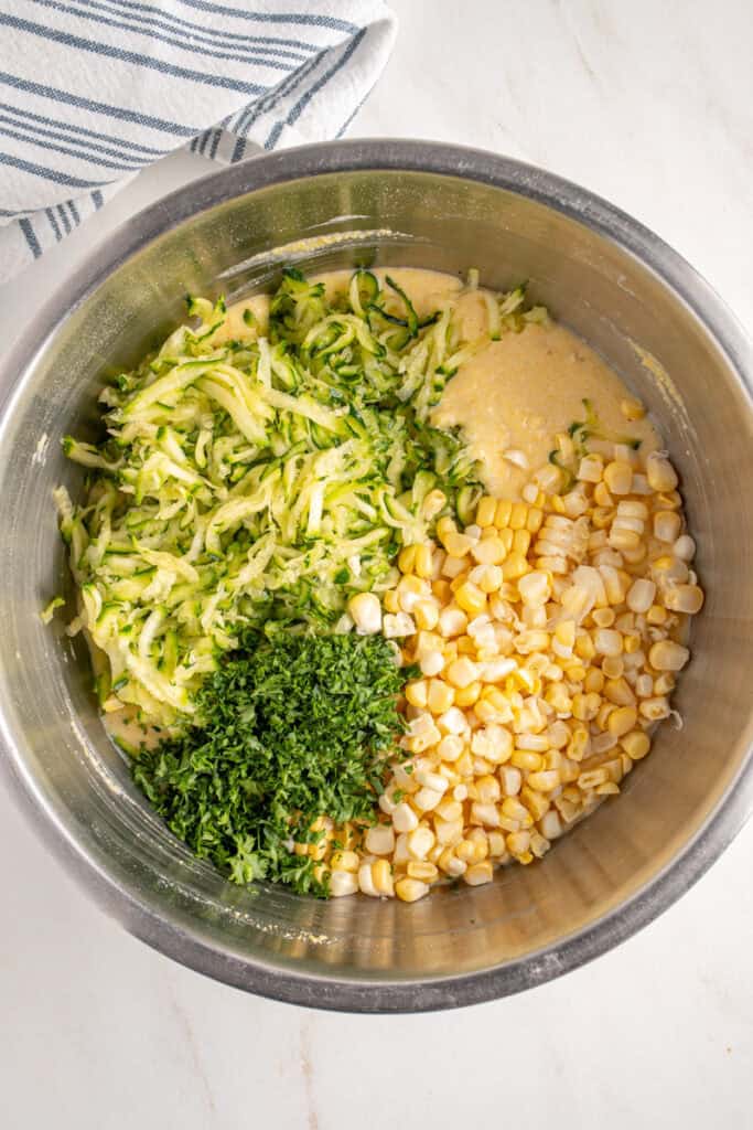 Adding corn, zucchini, and parsley to the corn meal batter in a bowl.