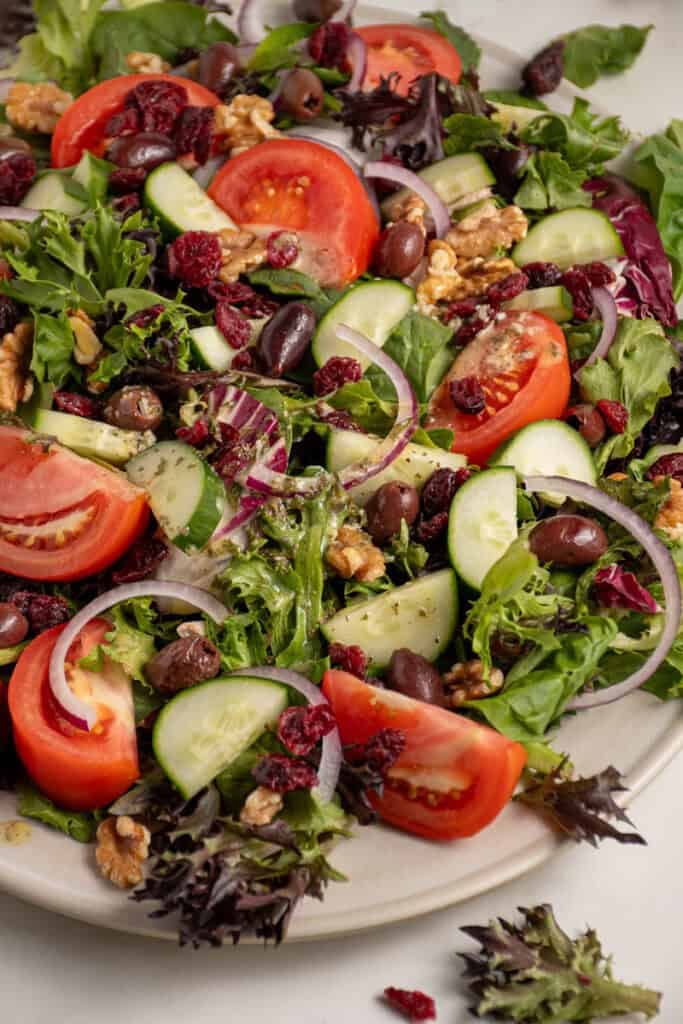 A large platter full of spring mix salad with tomatoes, cucumbers, red onion, walnuts, dried cranberries, and olives.