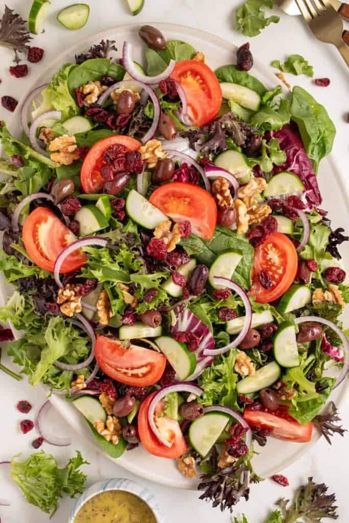 A large platter full of spring mix salad with tomatoes, cucumbers, red onion, walnuts, dried cranberries, and olives.