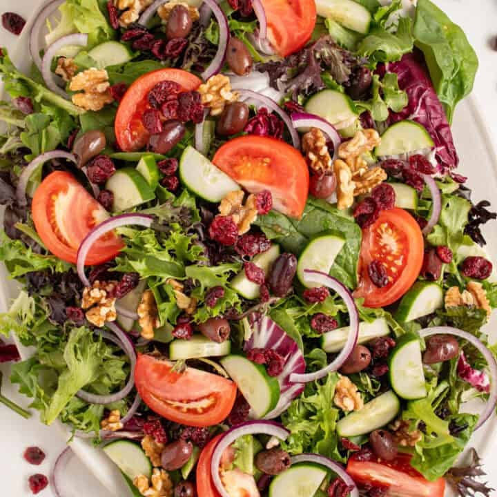 A large platter full of spring mix salad with tomatoes, cucumbers, red onion, walnuts, dried cranberries, and olives.