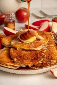 A drizzle of maple syrup being pored onto a stack of apple pancakes.