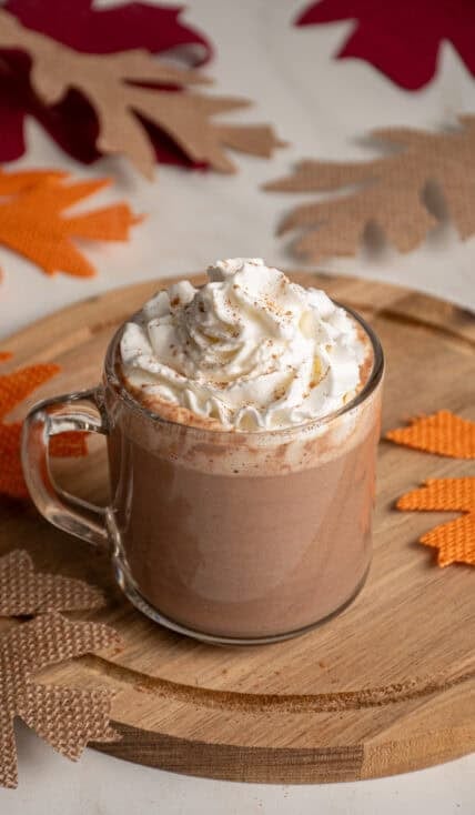 A clear glass mug with pumpkin hot chocolate and topped with whipped cream.